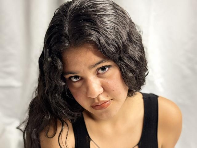 A photograph of CloverLover with wavy black hair, brown eyes, and a serious expression, wearing a black tank top, showing her bare shoulders and a slight pout.