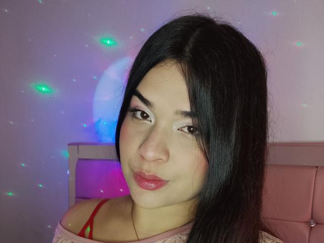 AntonelaPark has black hair, pink eyeshadow, red lipstick, and a cute smile, wearing a pink off-shoulder top, with a red strap visible, in a well-lit room.