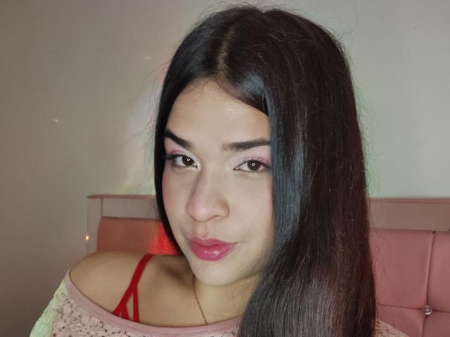 AntonelaPark has long dark hair, pink eyelid makeup, glossy pink lips, red bra straps, off-shoulder pink sweater, and a cute smile. Her ass is not visible.