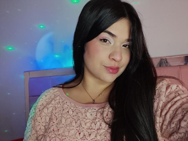AntonelaPark with long black hair, pink knitted sweater, pink lipstick, and a subtle smile, wearing a gold necklace, has green and blue fairy lights in the background.