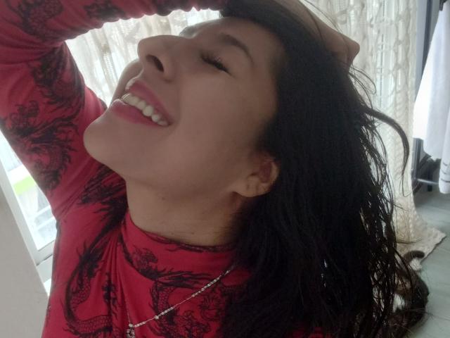 MalishkaFox with wet black hair, red dragon-patterned top, hand in hair, smiling, eyes closed, showing teeth, wearing a silver necklace, white curtains in background.