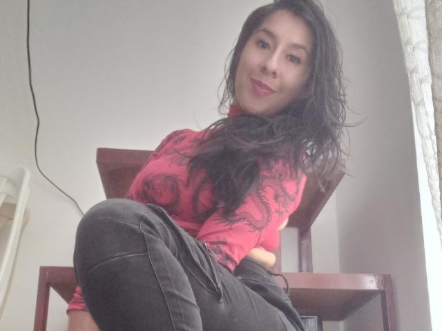 MalishkaFox sitting on a wooden chair, wearing a red, long-sleeve top with black patterns, and black pants, with long black hair and a slight smile, looking at the camera.