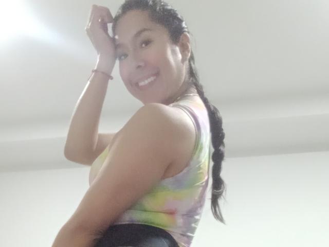 MalishkaFox with long braided hair, smiling, wearing a colorful tie-dye top, black pants, and a red bracelet, raising her right hand, showing her cute ass.