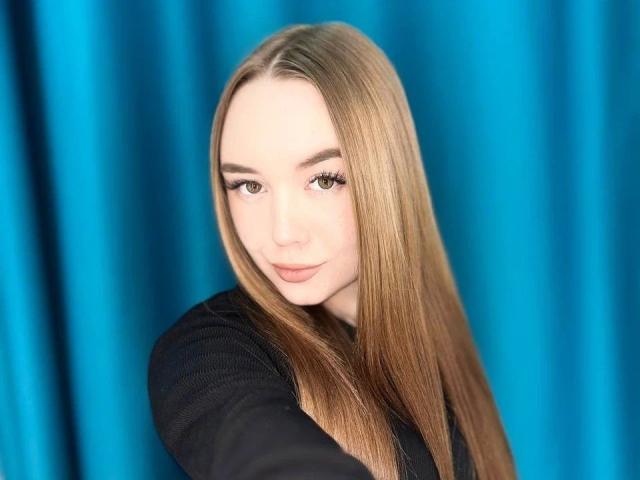 Marleee, de aspecto atractivo, con cabello rubio largo, vestida con una blusa negra, tiene pestañas prominentes y una sonrisa sutil, sus senos son ligeramente visibles, fondo de cortina azul, selfie, iluminación suave..