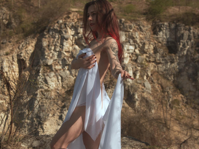 HotElements with red hair, holding a white sheet, exposing her ass and back tattoos, standing in front of rocky cliffs, sunlight highlighting her skin.