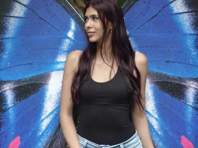 DanikaSerov with long red hair, black tank top, showing her small tits, and wearing blue jeans, standing against a blue, patterned wall. She has a cute, confident look.