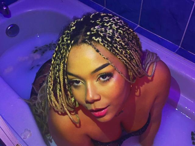 MarianaPrada with blonde braids, pink lipstick, and dark eyes, leaning in a white bathtub, wearing a black bra, looking up with a slight smile. Her tits are visible.