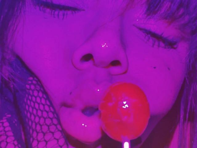 MarianaPrada's face, illuminated in purple, with eyes closed, a shiny red lollipop in her mouth, and fishnet covering her cheek. Her nose and lips are visible.