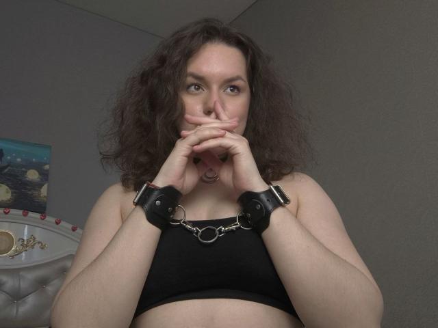 Dreia, con i capelli castani ricci, indossa un reggiseno nero, è manilata con manette nere, si morde un dito, ha un piercing al naso e guarda la telecamera con un'espressione leggermente seria..