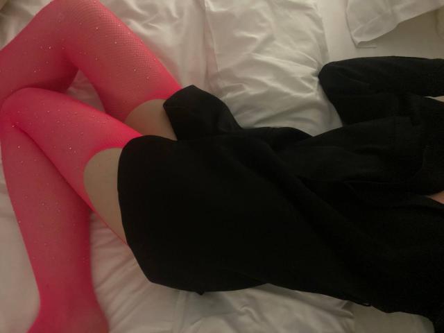 SofiButerfly lying on white bed, legs in pink glittery tights, black dress, butt barely covered, black top partially visible, soft lighting, skin visible above tights.