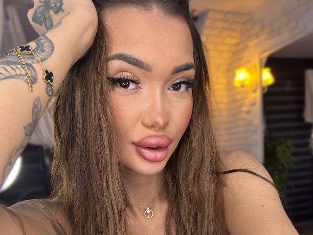 SashaFerrera heeft golvend bruin haar, volle, roze lippen, tatoeages op haar arm en dikke wimpers. Ze draagt een ketting en heft haar hand op. Zachte, warme verlichting..