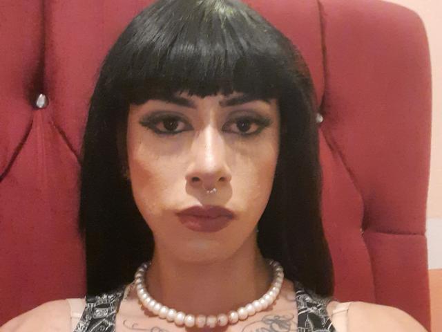 BusterHustler has black straight hair with bangs, a nose ring, thick eyeliner, and wears a pearl necklace. Visible tattoos on the chest. Red upholstered headrest.