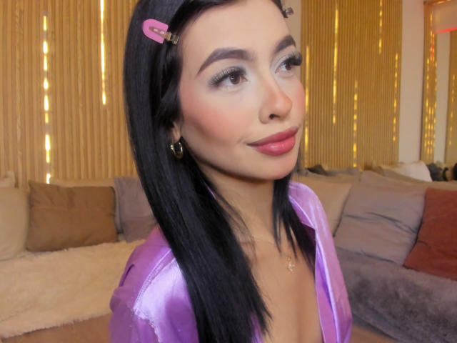 LeahMarie with black hair, pink hairclip, purple shiny top, pink lips, subtle makeup, and a slight smile, showing off her tits in a cute, sexy way.