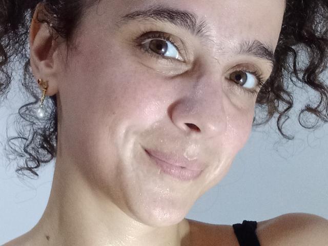 AmyFlores' face, showing curly brown hair, brown eyes, slight smile, small nose, and slightly shiny skin with visible pores, wearing a black strap, pearl earring on left.