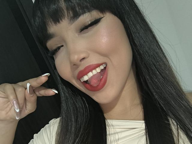 Heyly está lamiendo su lápiz labial rojo con la lengua fuera, los ojos medio cerrados, el pelo largo y negro, y las uñas blancas mejilla.