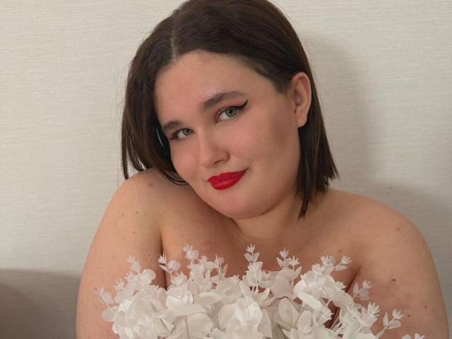 Jessica Joye, avec du rouge à lèvres rouge, un eye-liner noir et des cheveux bruns mi-longs, tenant des décorations florales blanches, souriant légèrement, montrant ses épaules dénudées et sa partie supérieure de la poitrine..