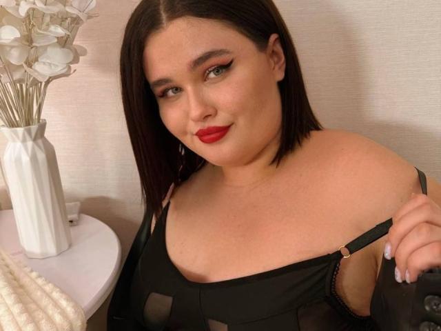 JessicaJoye with shoulder-length brown hair, red lipstick, and black sheer top, holding strap, exposing her tits, white vase with flowers on left, beige wall, smiling at camera.