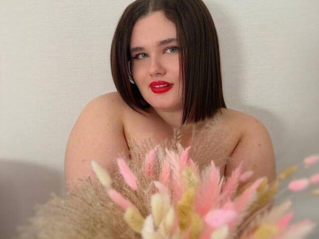 JessicaJoye con rossetto rosso, caschetto castano liscio e sfondo bianco, seduta con soffici cuscini rosa e beige davanti a lei, che mostrano le sue spalle scoperte e la parte superiore del petto..