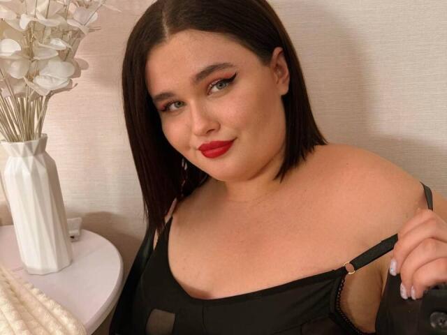 Jessica Joye, avec ses cheveux bruns foncés, son rouge à lèvres rouge et sa lingerie noire, tenant une bretelle, montrant ses épaules nues et sa poitrine supérieure. Vase blanc avec des fleurs sur la gauche..