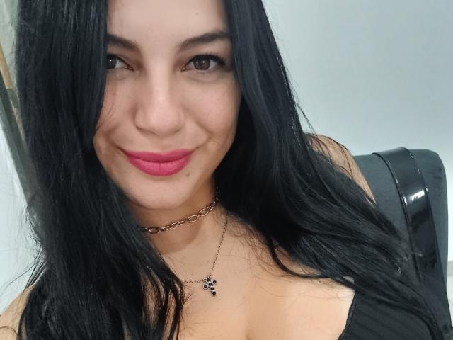 Paolitazz has long black hair, pink lipstick, and a slight smile. She's wearing a black top with a chain necklace and has a small pendant on her chest.