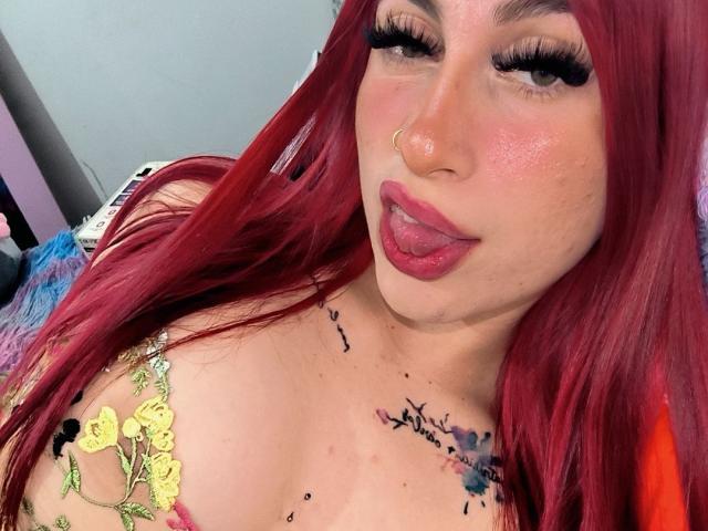 SammyViolet with red hair, nose ring, and long lashes, sticking out her tongue, showing her pink lipstick. She has tattoos on her chest and is wearing a floral top.