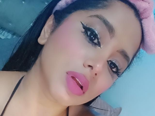 MariaCatalina's face with pink blush, black eyeliner, and pink lipstick, wearing a pink headband, looking at the camera with a slight open-mouth smile, showing her glossy lips.
