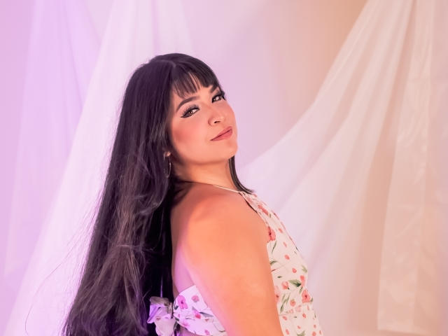 AntonelaPark with long black hair, floral dress, looking back with a slight smile, pink and white lighting, showcasing her tits and ass in the shot.