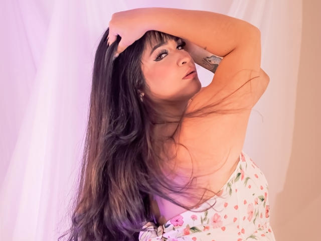 AntonelaPark with long brown hair, hands in hair, floral dress, looking at camera, pale skin, pink makeup, subtle tattoo on arm, soft pink light, slightly parted lips.