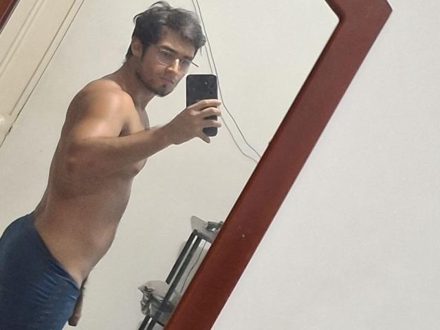 EliasX, shirtless with a toned, muscular build, wearing dark blue shorts, taking a mirror selfie with a black phone, showing his defined arm and slight facial stubble.