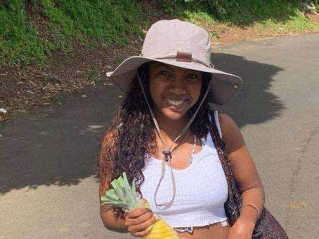 SexyValentinaLove with long curly black hair, tan skin, white crop top, and grey hat, holding a wad of cash and yellow fruit, smiling outdoors.