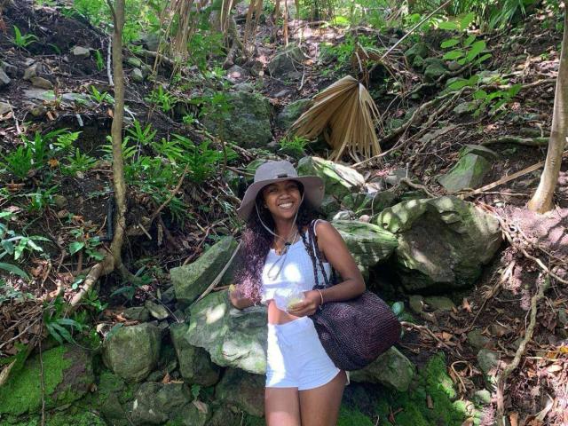 SexyValentinaLove standing in a forest, wearing a white tank top, brown hat, and brown backpack, smiling with long curly hair, green moss, and rocks behind her.