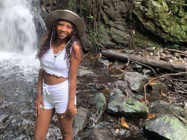 SexyValentinaLove with curly brown hair, white crop top, white shorts, and sun hat, standing by a waterfall, smiling, rock and tree background, wet rocks, forest setting.