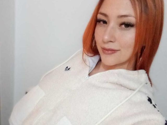 SamanthaCollen with orange hair, wearing a white hoodie, has a subtle smile, full lips, and hoop earrings, showing off her tits and a cute face.
