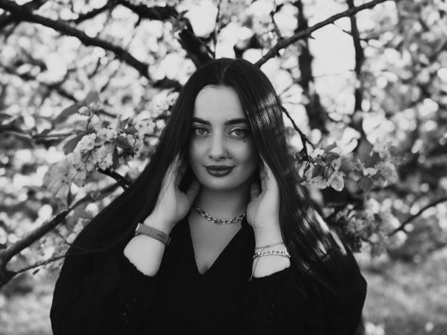 Black and white photo of MilanaPrice with long dark hair, hands in her hair, wearing a black top, necklace, and bracelets, smiling with dark lipstick, surrounded by blossoming branches.