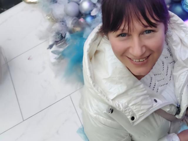 JasmineBlossom mit blauen Augen, lächelnd, trägt eine weiße, gepolsterte Jacke, ihr braunes Haar ist zu einem unordentlichen Dutt gebunden, sie sitzt auf einem weißen Fliesenboden, im Hintergrund sind blaue und weiße Ornamente zu sehen..