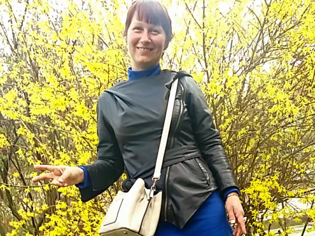 JasmineBlossom, con cabello castaño corto, sonriendo, vistiendo una chaqueta de cuero negra sobre un bolso azul y blanco, fondo de árbol amarillo, guiñando el ojo con la mano derecha, busto de tamaño medio, linda..