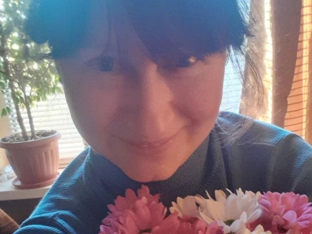 JasmineBlossom con fiori a forma di seno rosa e bianchi in primo piano, indossa un maglione blu, sorride leggermente, occhi azzurri, capelli castani, luce solare che filtra dalla finestra, pianta in vaso sullo sfondo..