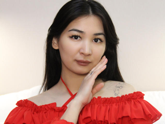 DarcyArnett with long black hair, wearing a red off-shoulder top, holding her chin with her right hand, showing a tattoo on her left shoulder. Cute, attractive Asian woman.