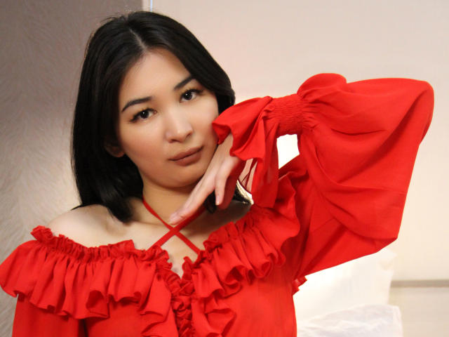 DarcyArnett with black hair, wearing a red off-shoulder top with ruffles, has her right hand near her chin, looking slightly to the side with a subtle smile.