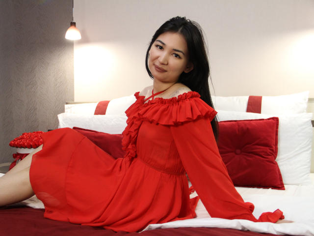 DarcyArnett with long black hair, red off-shoulder dress, sitting on bed with white and red pillows, smiling, looking at camera, red dress showing her tits, cute.
