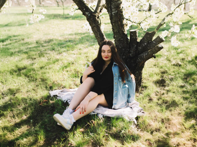 MilanaPrice, with pale skin and dark hair, sits on a blanket, wearing a black dress, denim jacket, and white sneakers, legs crossed, under a tree with white blossoms.
