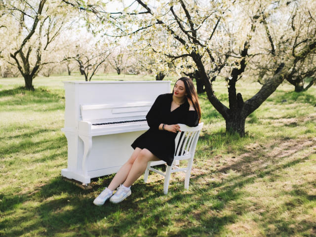 MilanaPrice, with pale skin and brown hair, sits on a white chair, arms resting on a white piano, wearing a black dress and white sneakers, in a grassy orchard.