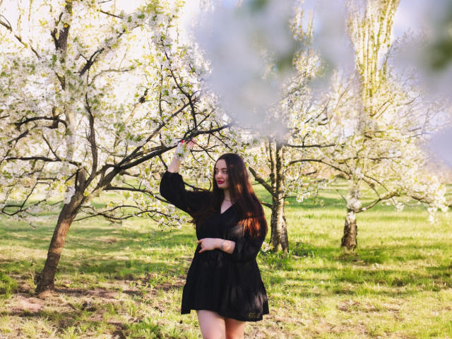 MilanaPrice with long brown hair, black dress, standing in grassy orchard, touching white-blossomed tree branch with right hand, smiling, sunlight, spring, green grass.