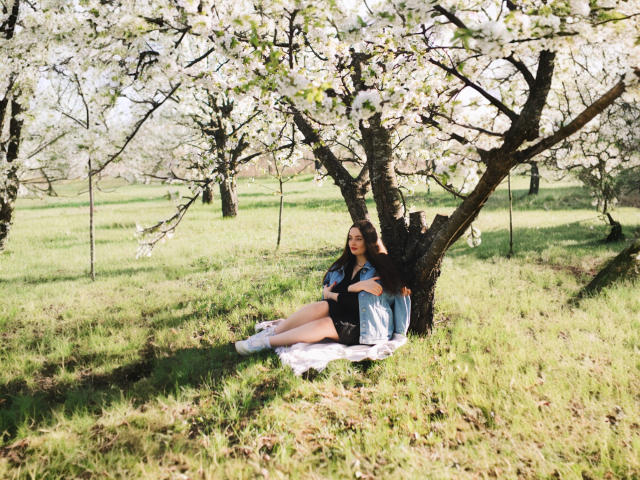 MilanaPrice is sitting against a tree with white blossoms, wearing a blue shirt, black shorts, and white socks, under a sunlit, green grassy area.