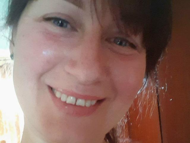 Il viso sorridente di JasmineBlossom, con un nasino carino, occhi azzurri e guance leggermente arrossate, i capelli castani e i denti bianchi, in un selfie informale..
