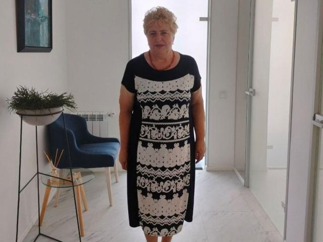 PannyGrannny è in una stanza, indossa un vestito a motivi bianchi e neri, ha i capelli biondi ricci e un sorriso, sullo sfondo si vedono una sedia blu e una pianta..