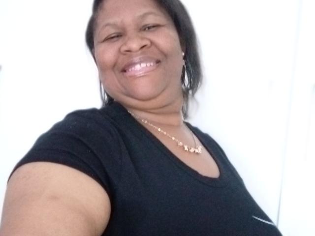 KillerBoobsLona, smiling with shiny pink lips, wearing a black shirt, gold necklace, and small earrings, has a medium build and dark skin, with short black hair.