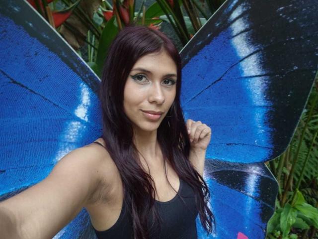 DanikaSerov with blue butterfly wings, black tank top, and long dark hair, smiling, holding her wing, showing her armpit, standing in front of green plants.