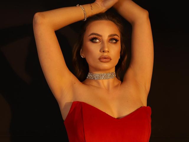 AllaLeo con le braccia alzate, indossa un abito rosso senza spalline, un collare di diamanti e un braccialetto, e guarda seducentemente l'obiettivo, con i capelli castani, un trucco scuro e un'espressione seria..