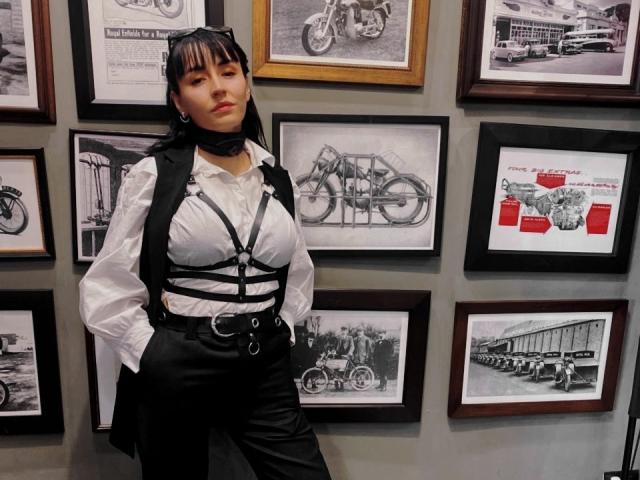 SaraleePoison stands with hands in black leather pants, white shirt, and black harness, showing her big tits and small ass. She has black hair and a serious face.
